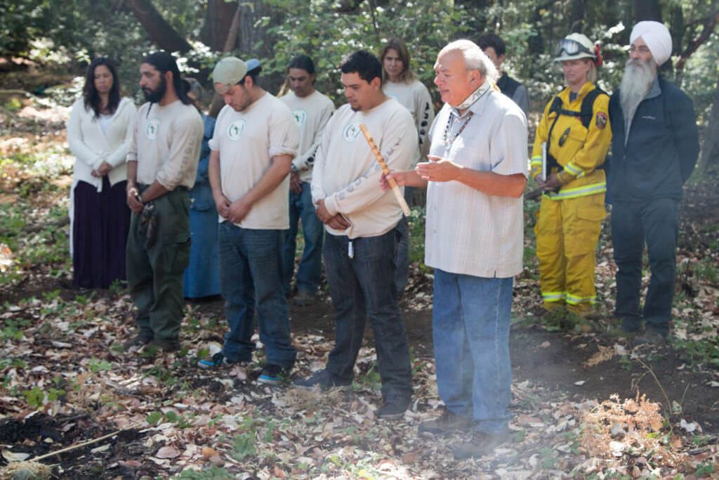 A faint cloud of smoke rises in front of his feet to his knees as Val Lopez, Chairman of the Amah Mutsun Tribal Band and President of Amah Mutsun Land Trust, leads a fire ceremony while tribal stand at his side and in a row behind them with eyes closed and heads bowed. A firefighter and additional people gathered for the prescribed burn to follow also look on from the row behind at San Vicente Redwoods, photo by Mike Kahn