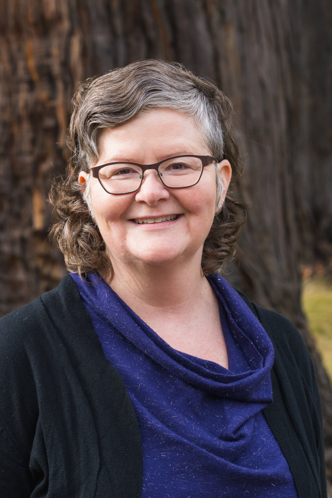 Claire SunSpiral smiles into the camera in front of a mature redwood tree, by Orenda Randuch