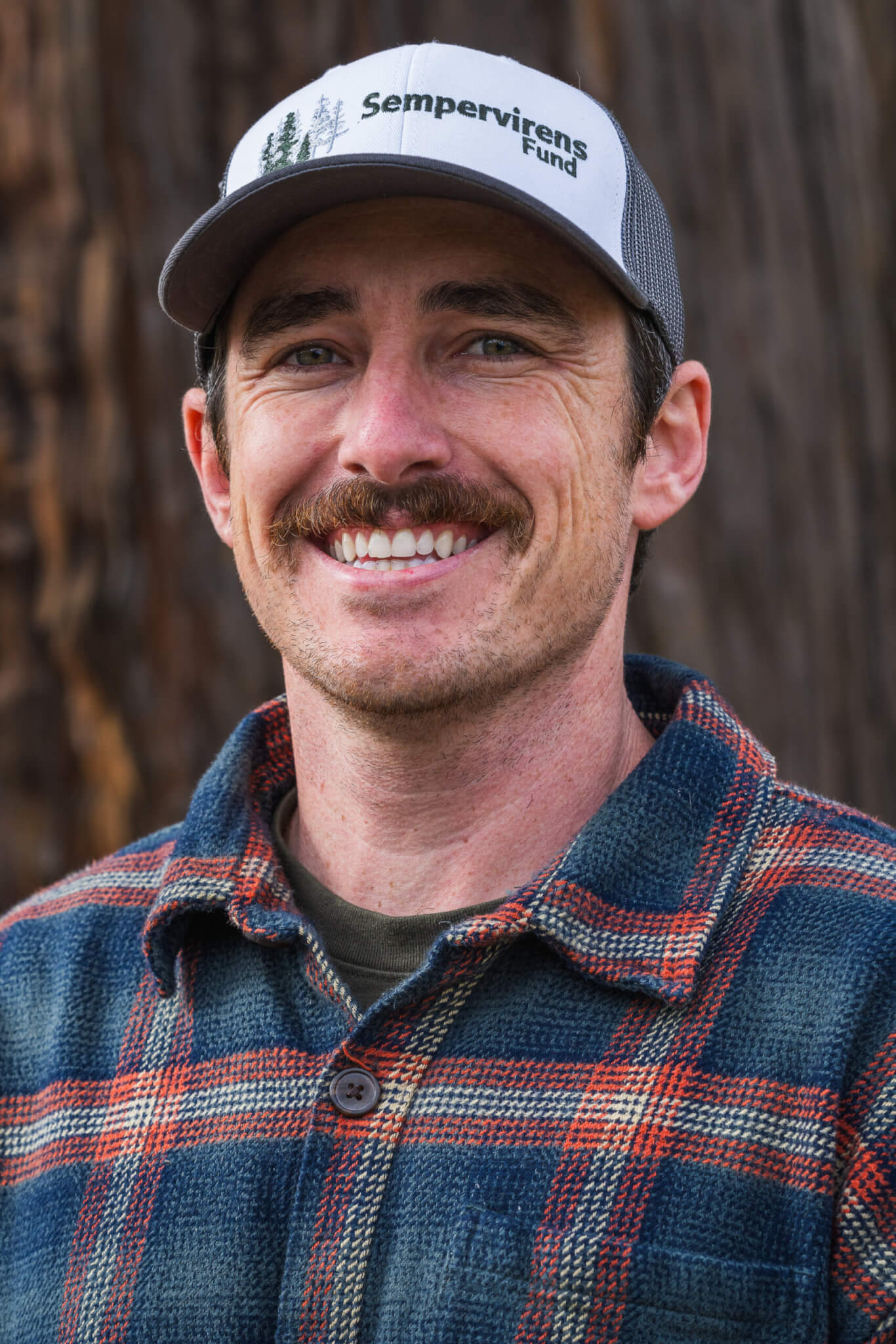 David Cowman smiles into the camera wearing a Sempervirens Fund hat in front of a mature redwood tree, by Orenda Randuch