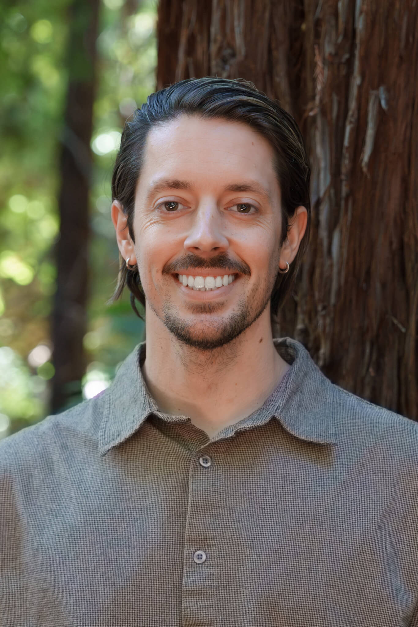 Robbie Brown smiles standing in front of a redwood tree in the forest, by Orenda Randuch