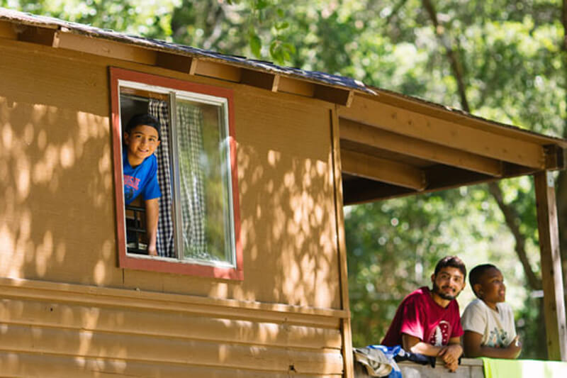 boy smiles out a cabin window and a counselor and another camper smile from the porch at Camp Jones Gulch