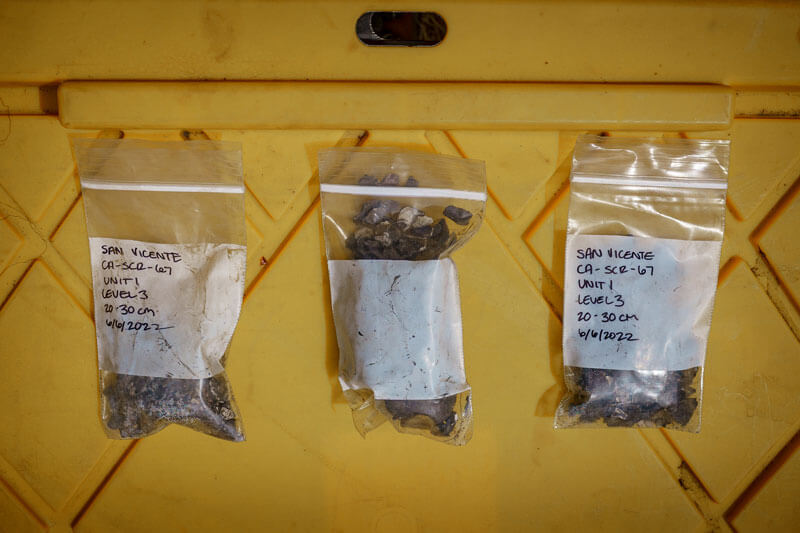 Three small plastic, labeled bags filled with tiny fragments of shell, bone, and other materials of various colors collected from soil samples lay against a bright yellow equipment box for later lab analysis, photo by Orenda Randuch