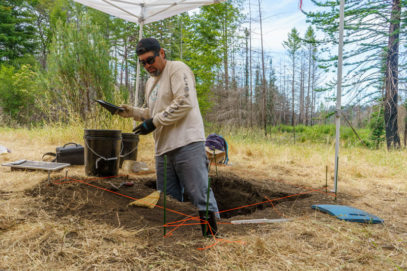 Lupe, Amah Mutsun Land Trust Native Steward, stands in an excavation plot up to his knees under the shade of a pop up tent as he measure soil samples a thin layer at a time in search of visible artifacts at San Vicente Redwoods where both green trees as well as recovering and dead trees can be seen behind him from the 2020 CZU Fire, photo by Orenda Randuch