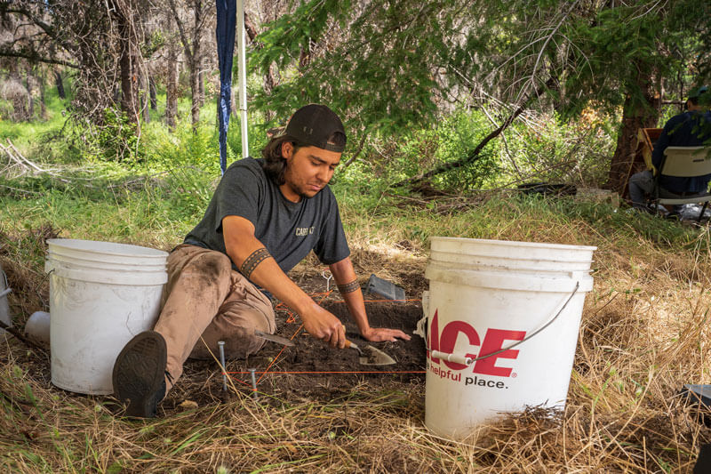 Nico, Amah Mutsun Land Trust Native Steward, sits on a dry grassy area between two white buckets using a small tool to carefully remove soil samples from a plot outlined by bright orange string at San Vicente Redwoods, photo by Orenda Randuch