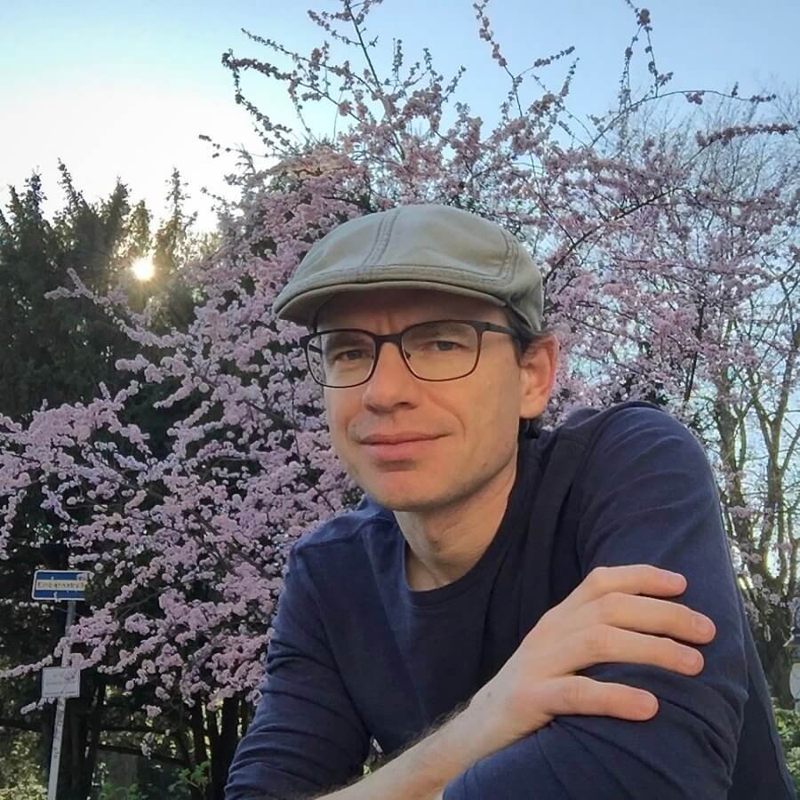 Author and historian Jared Farmer, dressed casually in a dark blue long-sleeved shirt with the sleeves pushed up, a light tan hat, and thin black framed glasses, smiles slightly from in front of a tree with light pink blooms and beyond the late afternoon sun peeks through a silhouetted tree under a soft blue sky