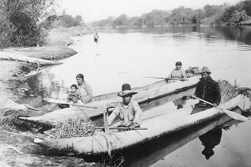 A historic black and white photo shows two canoe-like boast dug out of large tree trunks against a river bank where Indigenous Peoples sit toward the front and back of each boat.