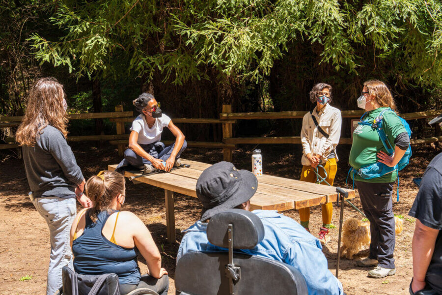 Disabled Hiker community members meet around a trailside accessible picnic table without benches in the way that works best for them - people utilizing wheelchairs approach the table, and several hikers stand, while one hiker sits on the top of the table, by Orenda Randuch