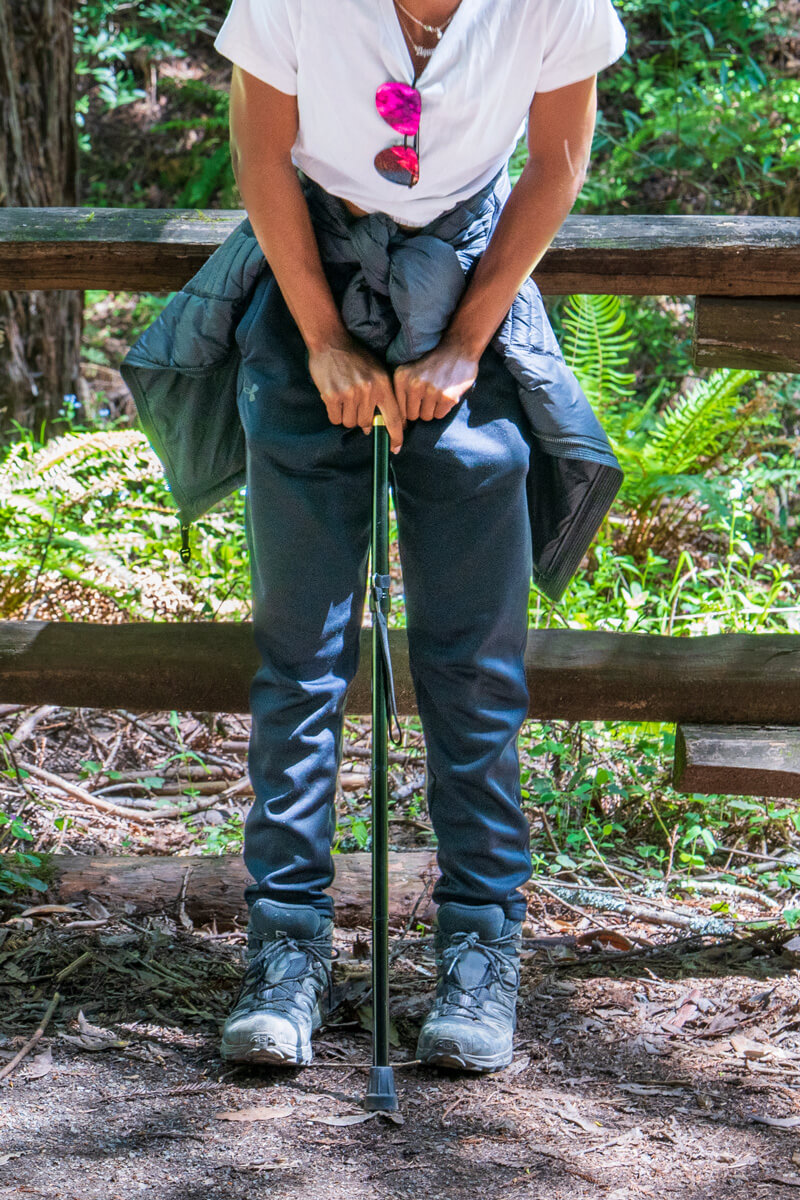 Visible from the shoulders down, a hiker with the Disabled Hikers community pushes down on an adjustable cane in front of two gray hiking boots along a rustic wooden fence and green ferns in the forest’s lush understory behind, by Orenda Randuch