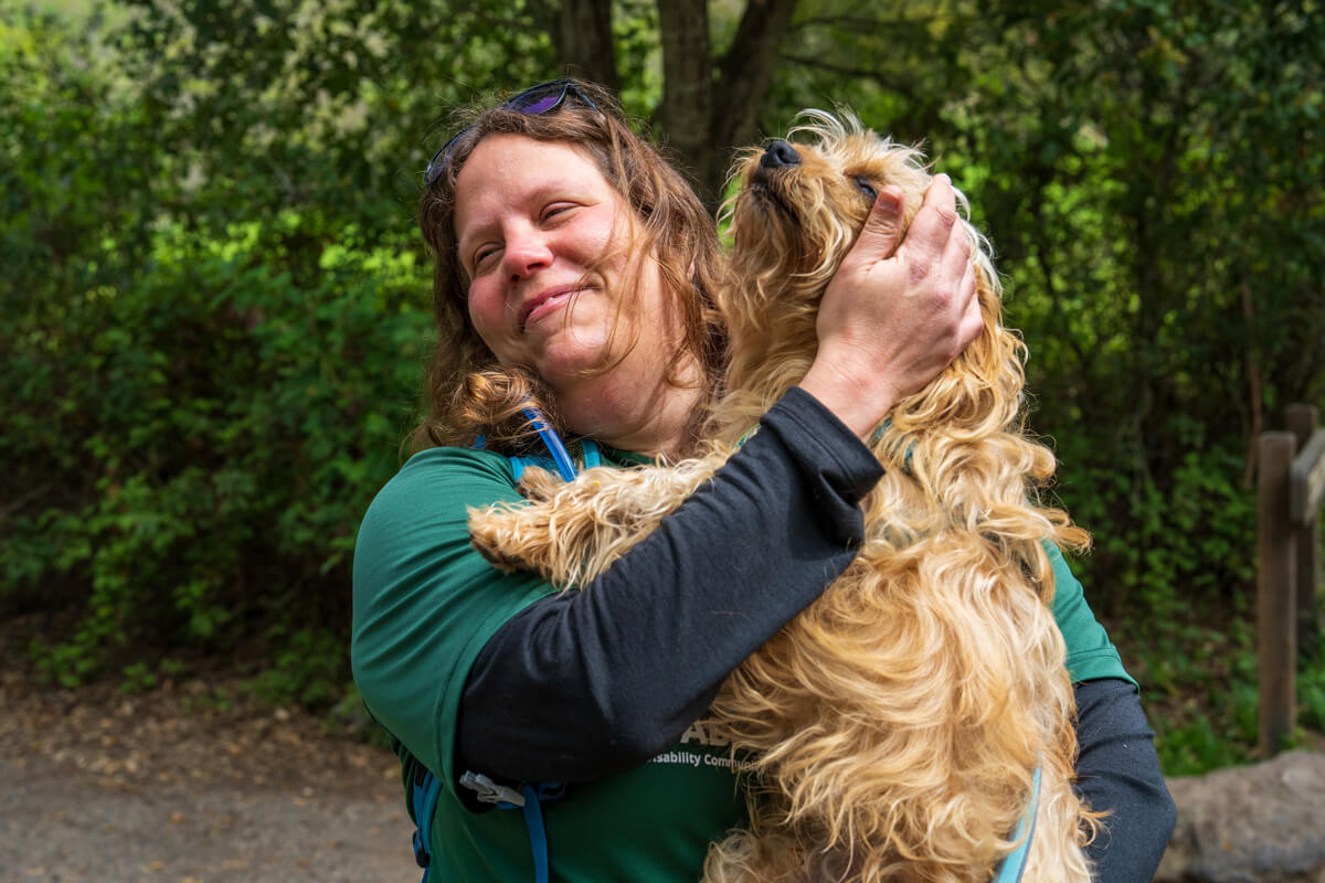Syren holds and pets her dog on the trail as both seem to smile blissfully looking to the left in front of a dark green forested backdrop, by Orenda Randuch