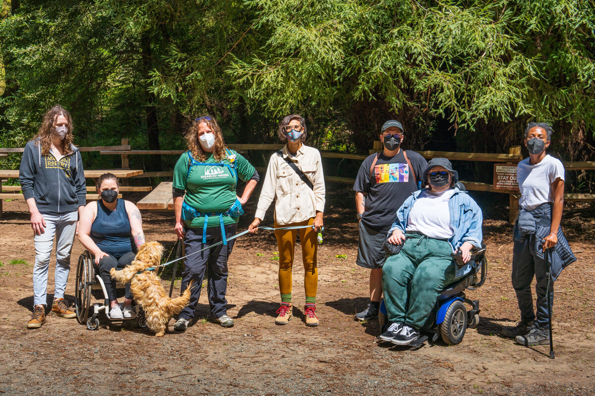 Seven members of Disabled Hikers donning face masks and hiking gear, which for some members include mobility devices like wheel chairs and canes, and a fluffy caramel colored dog on leash are lined up for a group photo in the sun with a lush redwood canopy behind them, by Orenda Randuch
