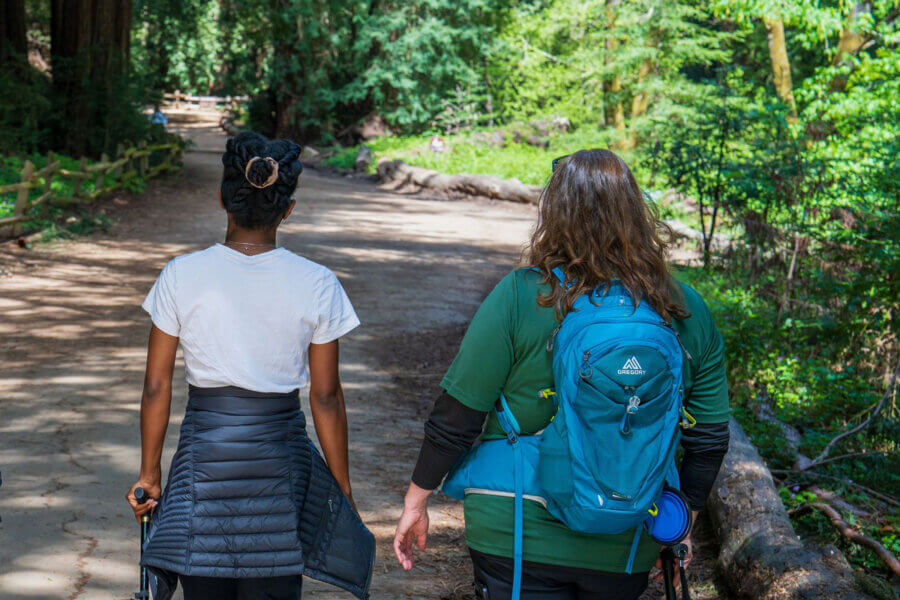 Syren and another member of the Disable Hikers community utilize canes on their hike along a trail that winds into the lush green redwood forest on a sunny day, by Orenda Randuch