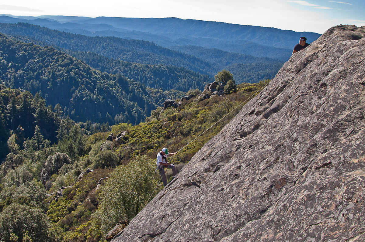 A belayer anchored behind the peak watches his climber repel back down the mottled gray and tan rock face high above the green forested slopes of the Santa Cruz mountains off into the horizon at Castle Rock State Park in late 2010 before the park put on the Governor’s list for closure, by Phillipe S. Cohen