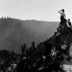 A black and white photo from Sempervirens Fund’s historic archive shows three men—in early 20th century suits consisting of dark colored trousers, white collared long-sleeved shirts with ties, dress shoes, and a few accessories like suspenders, vests, and hats reminiscent of fashion just after the turn of the century—sit casually atop a rock outcropping rising above the surrounding brush and forest like a spire as the view of forested mountainsides fade off into the distance behind them.