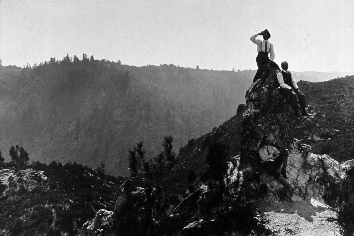 A black and white photo from Sempervirens Fund’s historic archive shows three men—in early 20th century suits consisting of dark colored trousers, white collared long-sleeved shirts with ties, dress shoes, and a few accessories like suspenders, vests, and hats reminiscent of fashion just after the turn of the century—sit casually atop a rock outcropping rising above the surrounding brush and forest like a spire as the view of forested mountainsides fade off into the distance behind them.