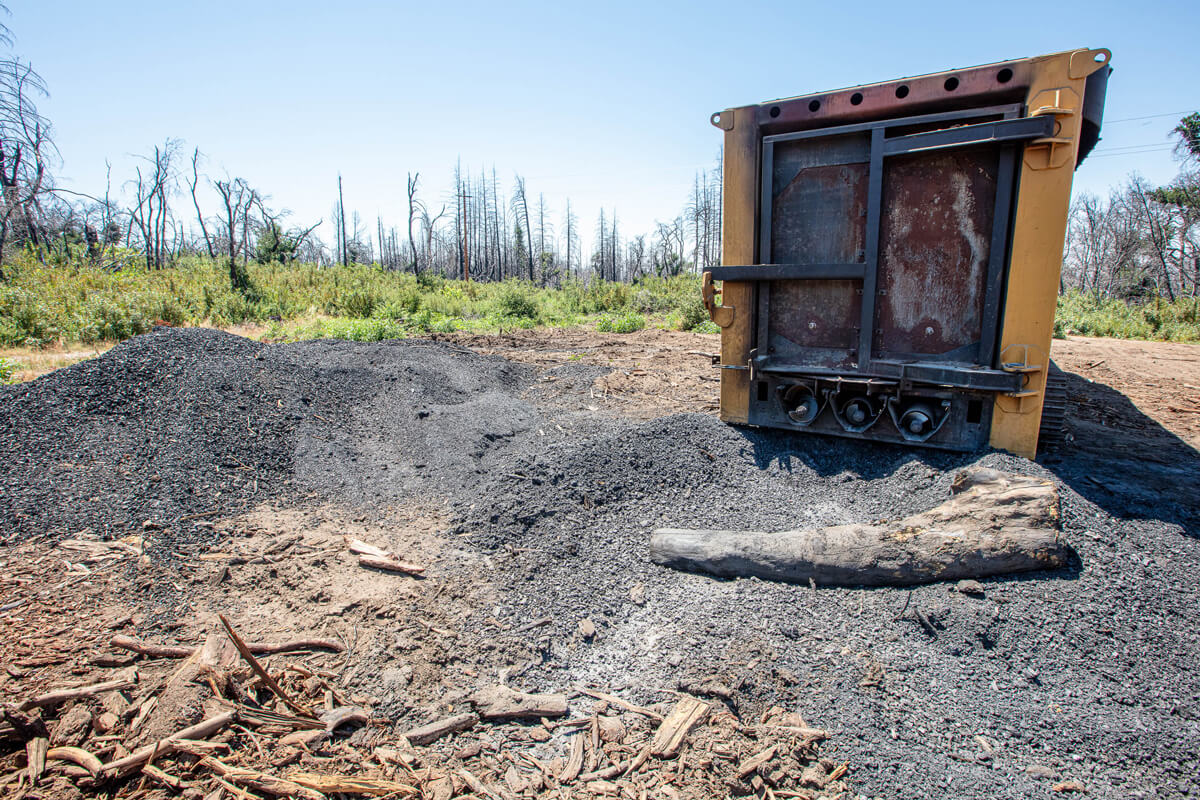 Resembling a yellow shipping container, a carbonator sits behind a large gray pile of bio char, the 10% left behind after burning off the bio mass of dead trees that can be used to return nutrients to the soil mimicking fire, by Ian Bornarth
