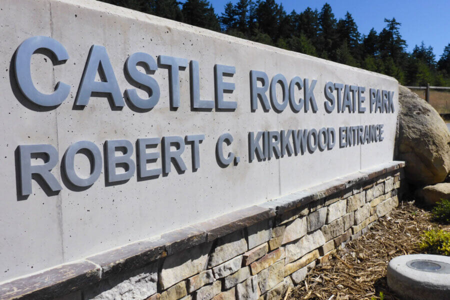 An up close view of the new Robert C. Kirkwood Entrance to Castle Rock State Park sign below the drak green treetops and bright blue sky beyond at the grand opening in 2019, photo by 7 Roots creative
