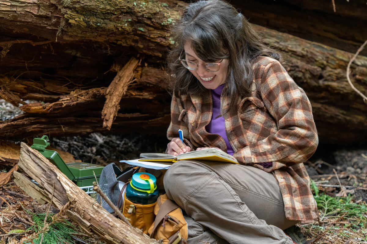 A person sitting on the ground near a redwood log smiles while writing in a notebook, by Orenda Randuch