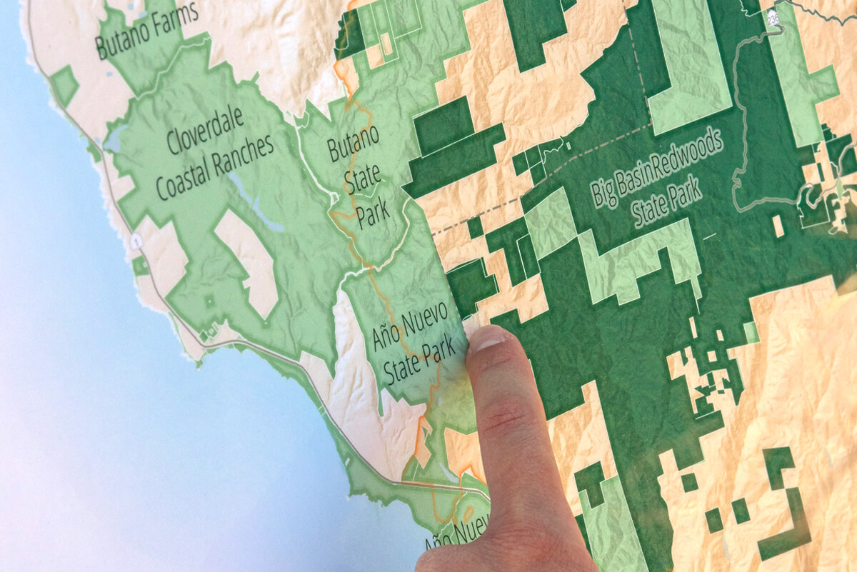 A finger points to the unprotected area on a map between Año Nuevo State Park, Big Basin Redwoods State Park, and Butano State Park, by Ian Bornarth