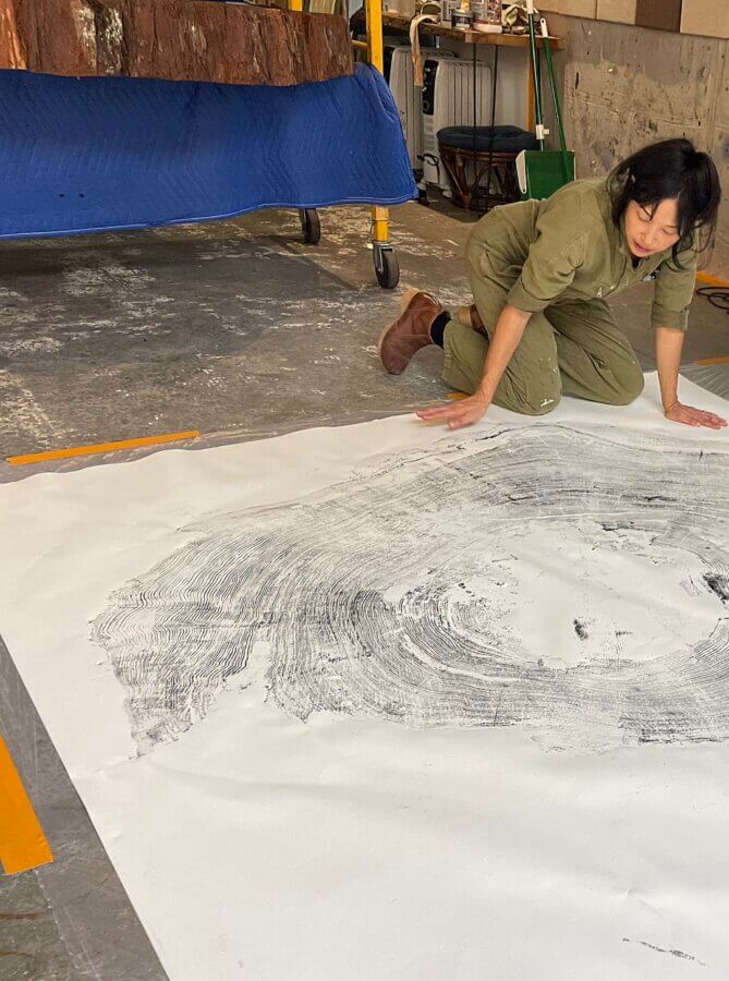 Artist Jane Kim kneels on the ground examining a print of redwood rings from a stump on a large piece of white paper, courtesy of Ink Dwell Studios