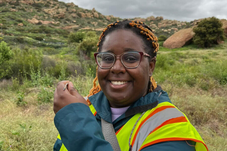 Dr. Earyn McGee smiles wearing a reflective safety vest and holding a small lizard in front of a shrub covered ridge, courtesy of Dr. Earyn McGee