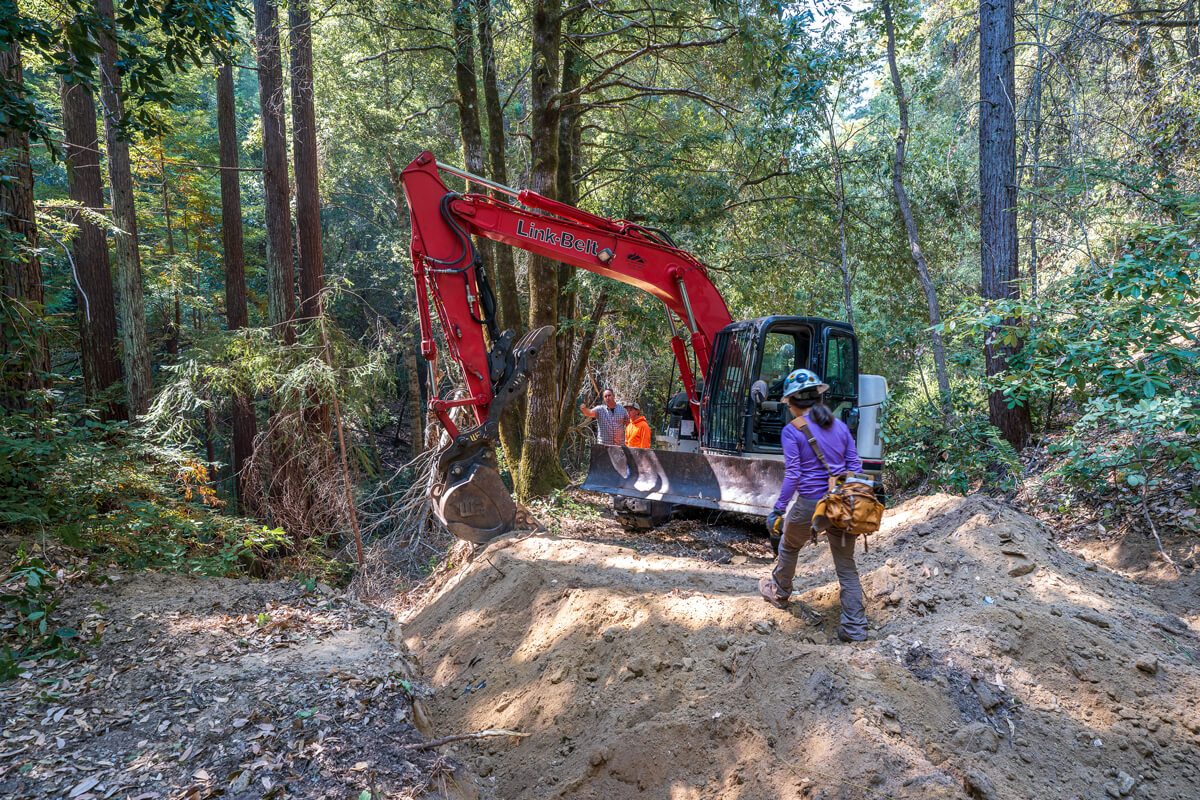 Crew in hard hats inspect soil and gravel from the unused road being moved back into a gentle slope along the creek bed by a tractor, by Orenda Randuch