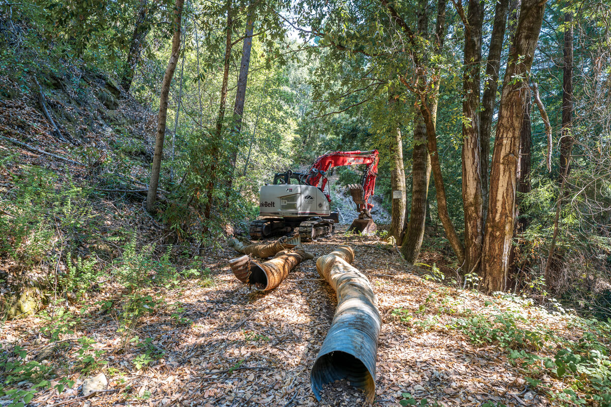 Two large worn and rusty corrugated metal pipes removed from the creek lay on the unused road behind a tractor that unearthed them, by Orenda Randuch