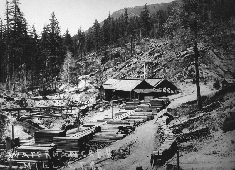 A black and white logged hillside behind a wooden mill surrounded by stacks of lumber, railroad tracks, and logging roads, with “Waterman Creek Mill C.T.C.” written in the bottom left, Sempervirens Funds Historic archive