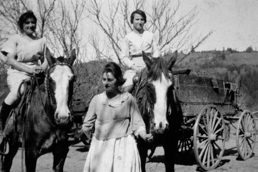 Three people pose outdoors; two are sitting on horses while one stands between them, holding the horses’ bridles. A wooden wagon and leafless trees are in the background, with hills in the distance. The photo is black and white.