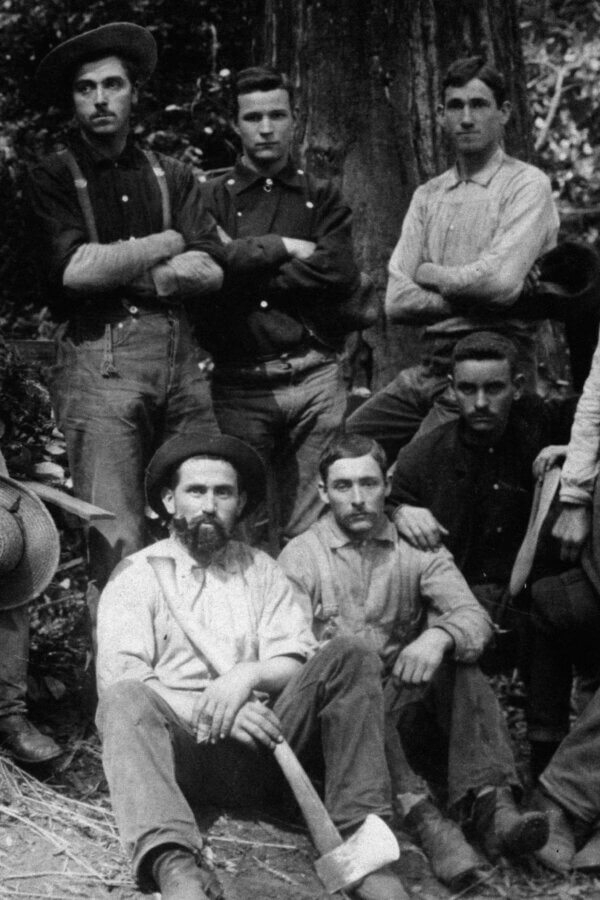 A black-and-white photo of six people in work clothes, some sitting, some standing, outdoors in a wooded area. One seated person holds an axe.