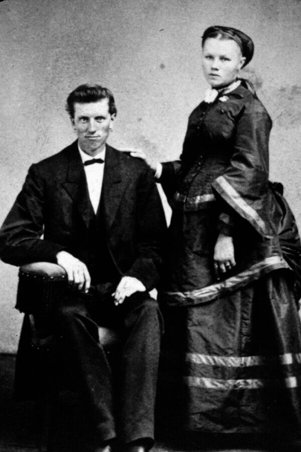 A black-and-white vintage photograph of a seated person in a suit and bowtie next to a standing person in a long, formal dress with a bonnet, both posing for a formal portrait against a plain backdrop.