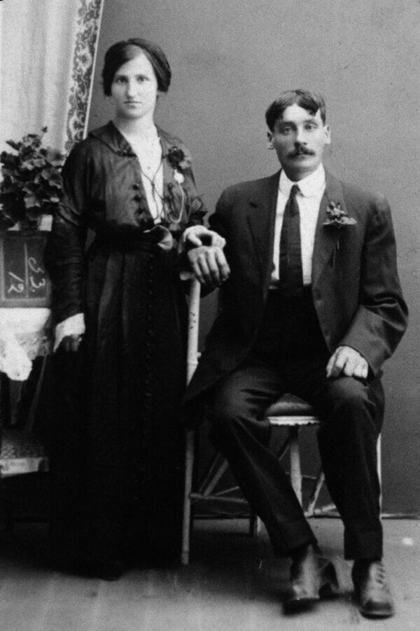 A black and white vintage photograph of a person in a long dark dress standing beside a seated person in a suit and tie. They both look serious, posing for a formal portrait with one hand resting on his arm.