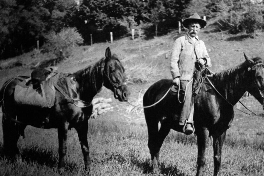 A person wearing a hat and suit sits on a horse, holding the reins of a second saddled horse beside him, with grassy ground and trees in the background. The photo is black and white.
