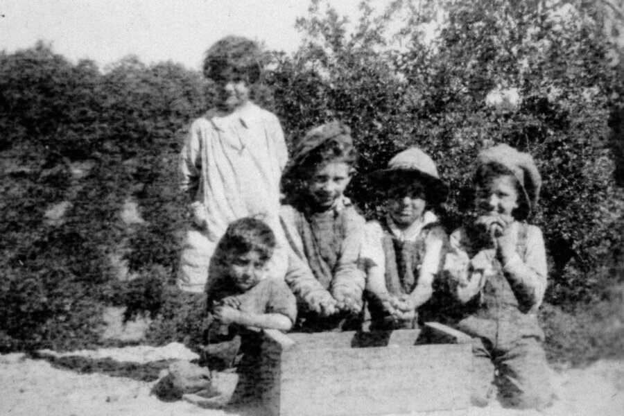 Five children pose outdoors in old-fashioned clothing, with greenery in the background. Four children kneel or sit around a wooden box, while one stands slightly behind them. The image is black and white and appears vintage.
