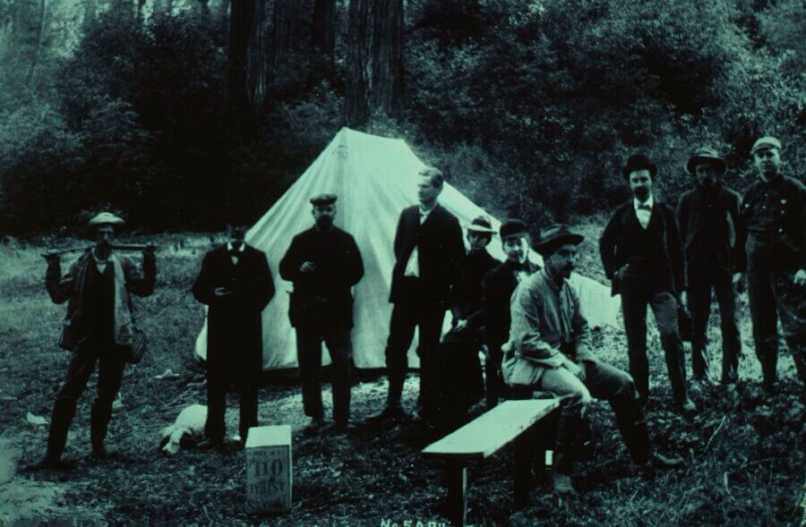 A group of people in old-fashioned clothing stands and sits in front of a canvas tent in a wooded area, suggesting a historical camping scene. Some wear hats and suits; trees and foliage surround the camp.