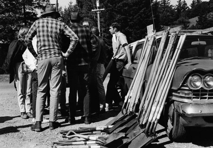 A group of people stand in a circle on a dirt road, seen from behind. Nearby, a car is parked with shovels and tools leaning against it. Trees and power lines are visible in the background.