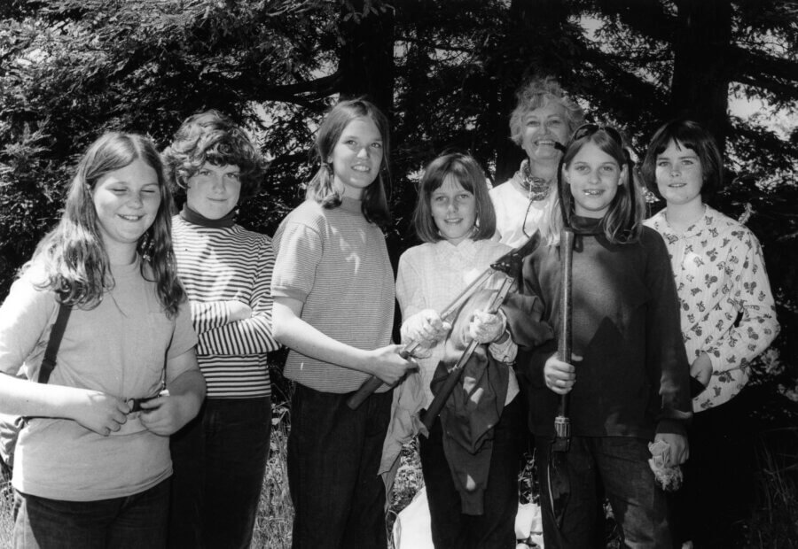 A group of six people stand outdoors in front of trees, smiling at the camera. Some hold hiking sticks and outdoor gear, suggesting they are on a nature outing.