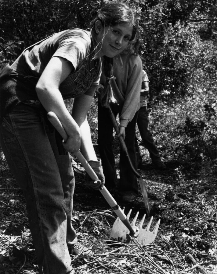 A young person outdoors uses a rake to clear debris while other people work in the background in a natural, wooded area.