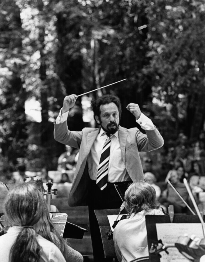 A conductor in a suit and striped tie leads an orchestra outdoors, raising a baton with both hands. Musicians are visible in the foreground, and a wooded audience area appears in the background.