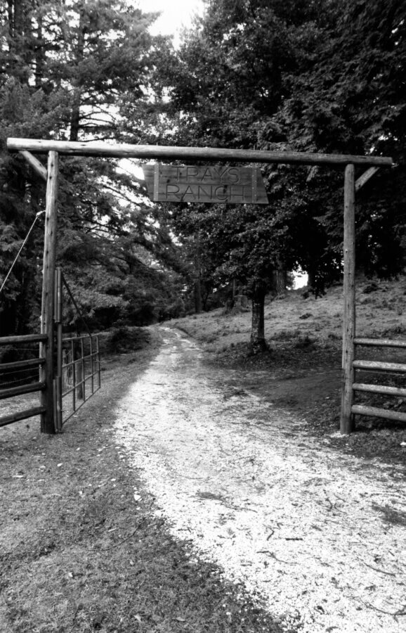 A black-and-white photo of a rustic wooden entrance gate labeled “RANCH,” leading to a gravel path surrounded by trees and fencing in a rural setting.