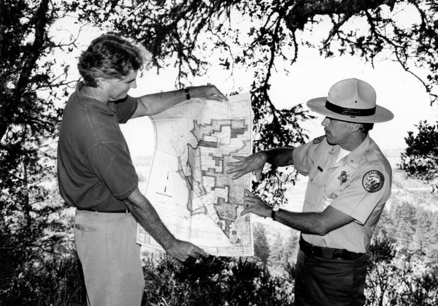 Two people stand outdoors under tree branches, holding and examining a large map. One person wears a park ranger uniform and hat, while the other is casually dressed. Both point to areas on the map, discussing its details.