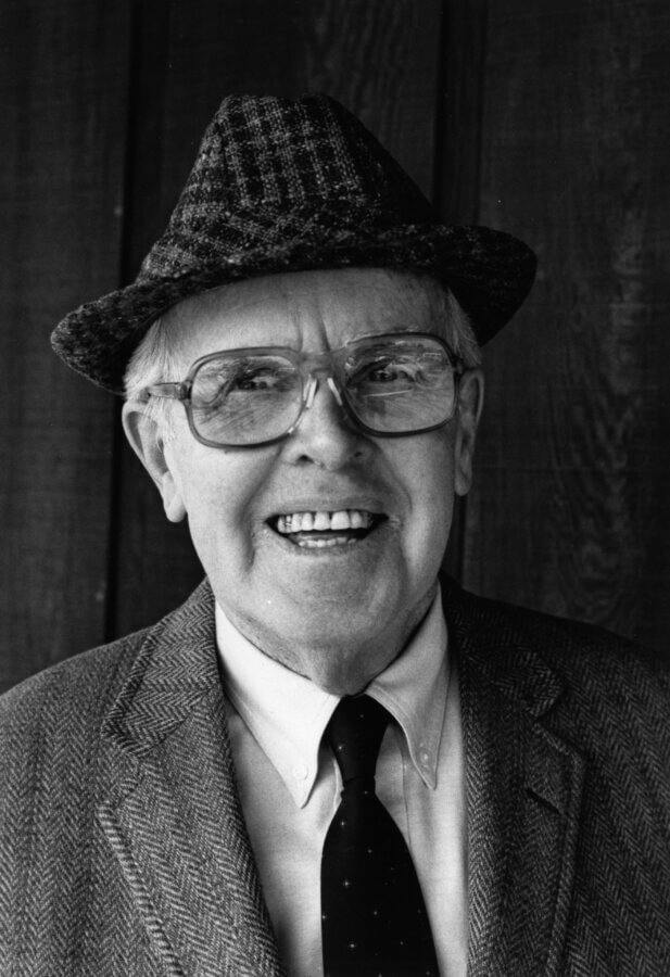 An person wearing glasses, a patterned hat, and a suit jacket with a tie smiles warmly at the camera. The background is a dark wooden wall.