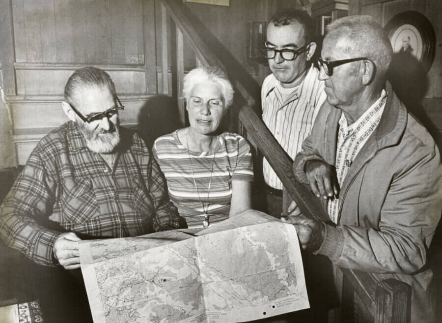 Four people look at a large map spread out on a table. They are indoors, standing close together, appearing to discuss or examine the map intently.