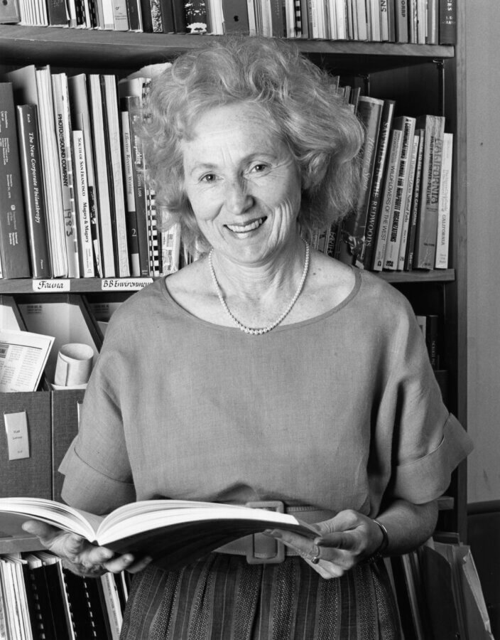 A person with curly hair and a pearl necklace smiles while holding an open book in front of a bookshelf filled with various books and files.