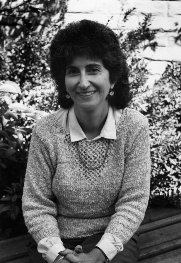 A person with curly dark hair, wearing a light-colored sweater over a collared shirt, sits on a bench outdoors, smiling at the camera with greenery and a brick wall in the background.