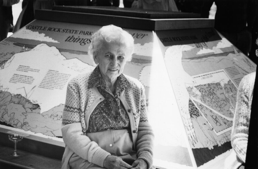 A person with short white hair sits in front of an illustrated display at Castle Rock State Park, wearing a patterned shirt, cardigan, and skirt.