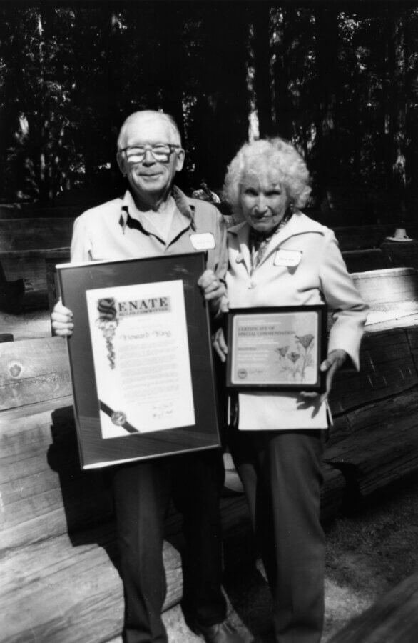 Two people stand outdoors holding framed awards. One wears glasses and smiles, holding a large Senate certificate. One has curly hair and holds a smaller framed document. They stand in front of wooden benches.