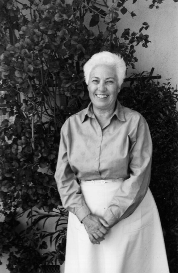 A person with short white hair smiles warmly, standing in front of leafy plants. The photo is black and white.