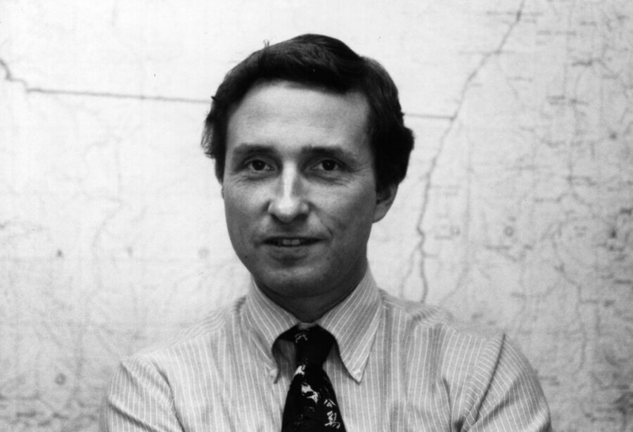 Black and white photo of a person with short dark hair, wearing a striped shirt and patterned tie, standing in front of a map on the wall.