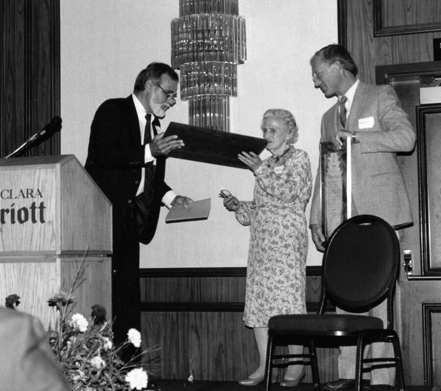 Three people stand together at an event. One person hands a plaque to another person, who is smiling.