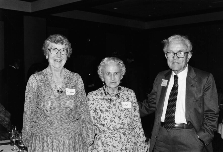 Three people smile at an outdoor event with other people in the background. All three wear name tags. Trees and a bright sky can be seen behind them.